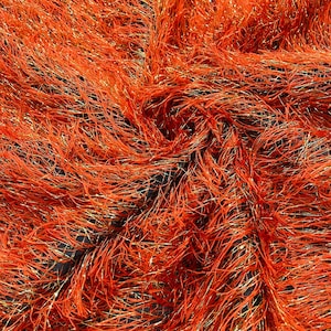 Puede incluir: Primer plano de una tela naranja texturizada. El material está compuesto por numerosos hilos finos, algunos con brillo dorado metálico, creando una superficie densa y táctil. El color de la tela es un tono naranja cálido y acogedor.