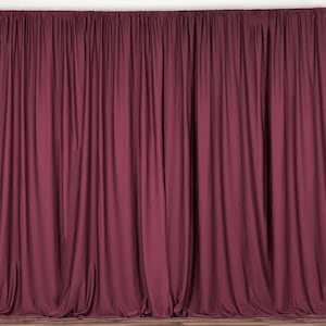 May include: A full shot of a burgundy curtain backdrop. The fabric is gathered at the top and hangs in vertical folds. The curtain is a solid colour and is set against a white wall and a wooden floor.