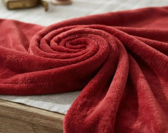 Puede incluir: Una manta de forro polar de color rojo intenso con una textura suave y esponjosa. La tela está dispuesta en un pliegue en espiral central, mostrando el pelo denso y aterciopelado del material. La manta muestra un color carmesí sólido.