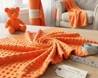 Puede incluir: Una tela minky de puntos color naranja neón con textura en relieve, acompañada de un oso de peluche a juego y un rollo de tela. Una cinta métrica muestra un ancho de 147,32 cm y una etiqueta dice Neon Orange Minky Dot Fabric - 58" Wide.