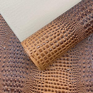 May include: A roll of brown faux leather material featuring a raised, embossed crocodile skin texture. The surface displays a repeating pattern of glossy, dark brown scales against a lighter brown base, with a light beige fabric backing visible.