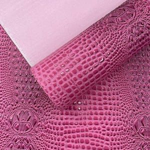Peut inclure: Gros plan sur un rouleau de similicuir rose avec une texture peau de crocodile. Le matériau est d'une nuance vibrante de fuchsia, avec une finition brillante. Un tissu rose plus clair est visible en arrière-plan.