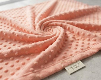 Puede incluir: Una tela minky de color coral con un patrón de puntos en relieve por toda la superficie. Una etiqueta de tela blanca está unida a la esquina, con el texto impreso: CORAL MINKY DOT 58" WIDE.
