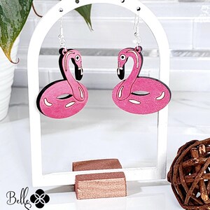 May include: Pink flamingo earrings with black accents and silver hooks. The earrings are displayed on a white arch-shaped stand. The earrings have a pink body and a black outline. The earrings are a fun, tropical accessory.