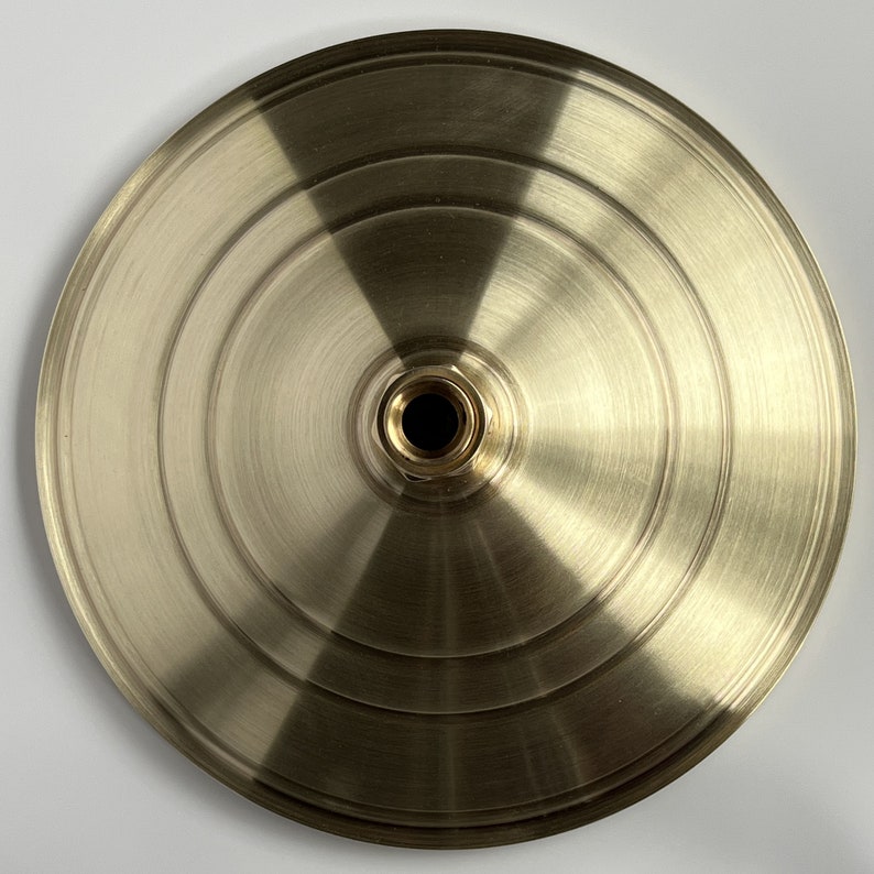 Puede incluir: Una placa de techo redonda de metal dorado con un agujero central. La placa tiene un acabado cepillado y un borde ligeramente elevado.