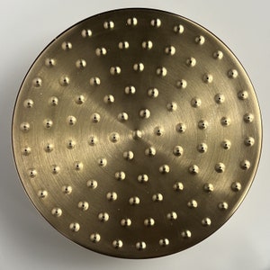 Puede incluir: Un cabezal de ducha redondo de color dorado con una superficie texturizada y m&uacute;ltiples agujeros peque&ntilde;os.