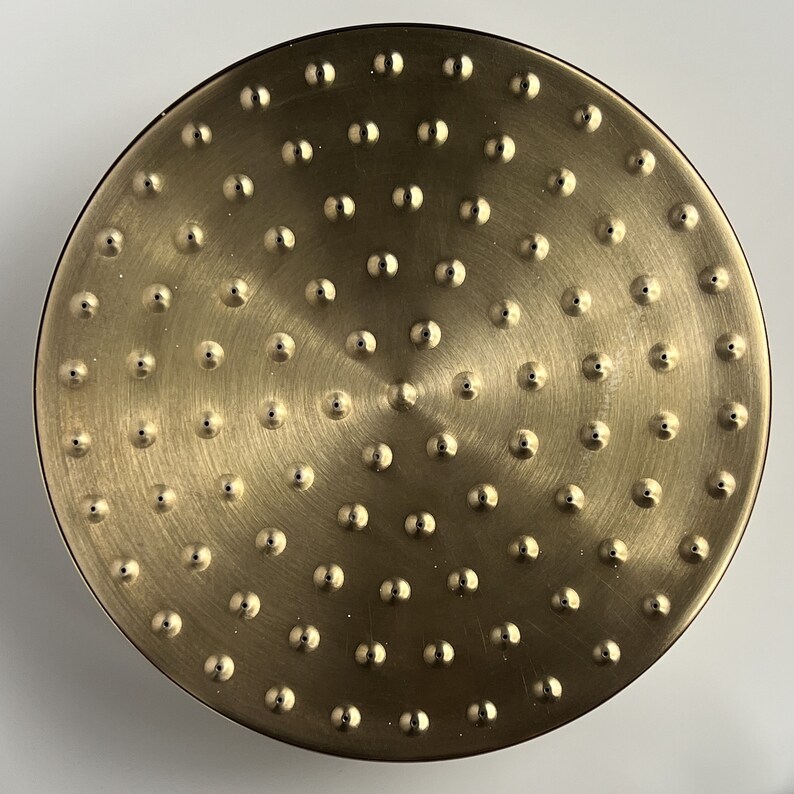 Puede incluir: Un cabezal de ducha redondo de color dorado con una superficie texturizada y m&uacute;ltiples agujeros peque&ntilde;os.