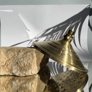 Puede incluir: Un cabezal de ducha dorado con una base redonda y plana y una parte superior abovedada. El cabezal de ducha est&aacute; sobre una roca marr&oacute;n y rugosa.