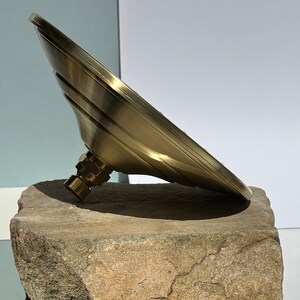 Puede incluir: Un cabezal de ducha de color dorado con un dise&ntilde;o redondo y plano y una superficie texturizada. El cabezal de ducha est&aacute; apoyado sobre una roca marr&oacute;n rugosa.