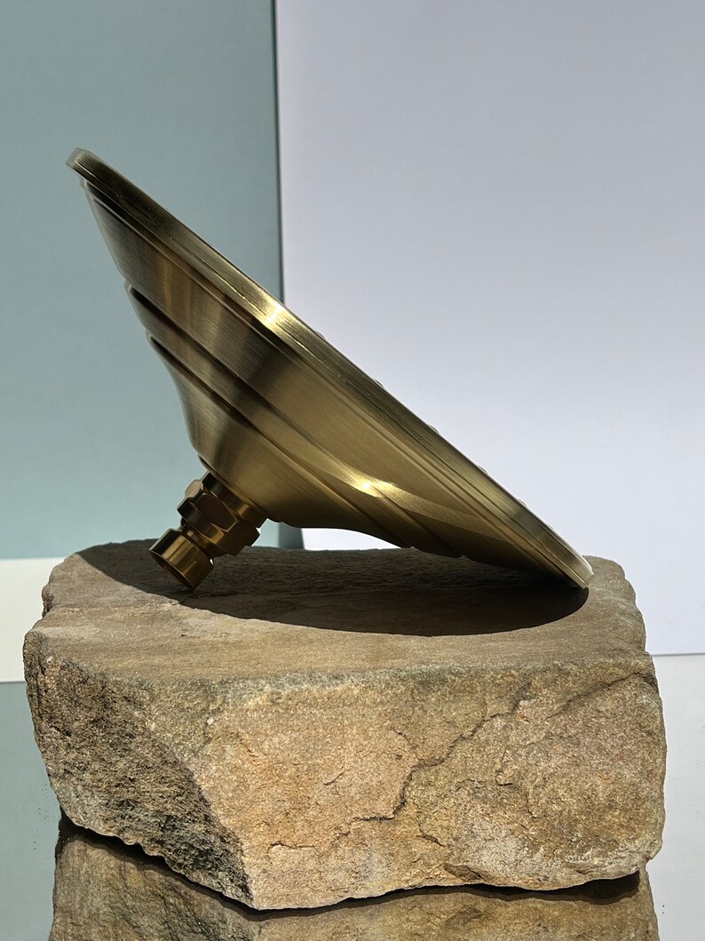 Puede incluir: Un cabezal de ducha de color dorado con un dise&ntilde;o redondo y plano y una superficie texturizada. El cabezal de ducha est&aacute; apoyado sobre una roca marr&oacute;n rugosa.