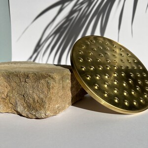 Puede incluir: Un cabezal de ducha de metal dorado con un dise&ntilde;o circular y una superficie texturizada. El cabezal de ducha est&aacute; sobre una roca marr&oacute;n claro.