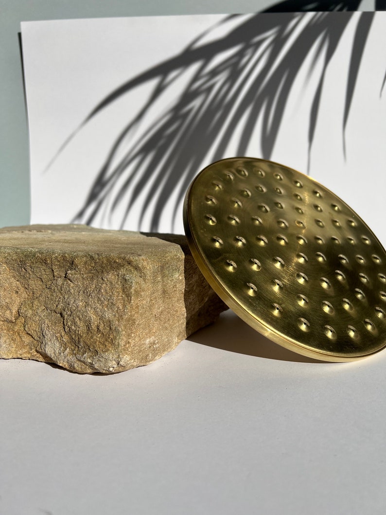 Puede incluir: Un cabezal de ducha de metal dorado con un dise&ntilde;o circular y una superficie texturizada. El cabezal de ducha est&aacute; sobre una roca marr&oacute;n claro.