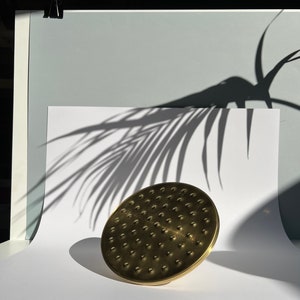 Puede incluir: Un cabezal de ducha dorado con un dise&ntilde;o circular y una superficie texturizada. El cabezal de ducha est&aacute; sobre una superficie blanca con la sombra de una hoja de palma.