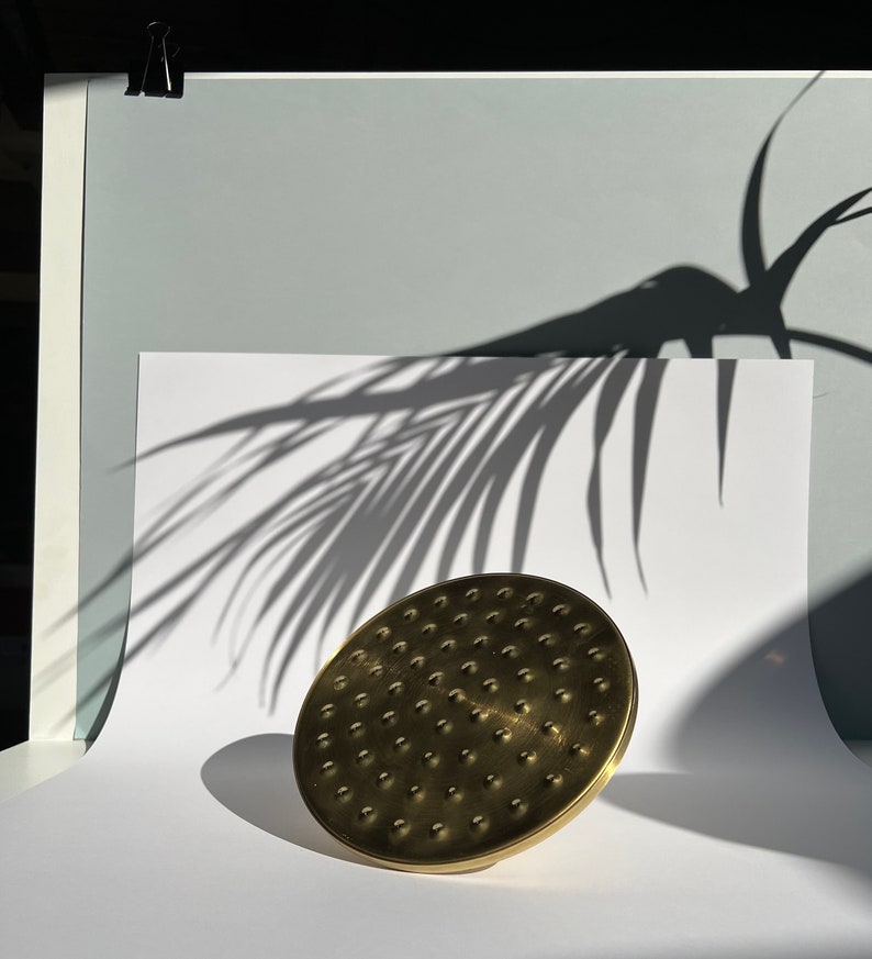 Puede incluir: Un cabezal de ducha dorado con un dise&ntilde;o circular y una superficie texturizada. El cabezal de ducha est&aacute; sobre una superficie blanca con la sombra de una hoja de palma.