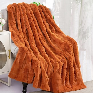 May include: An orange faux fur throw blanket draped over a gray armchair. The blanket has a textured, ribbed design and a soft, plush appearance. The chair is positioned next to a white cabinet with a decorative floral arrangement.