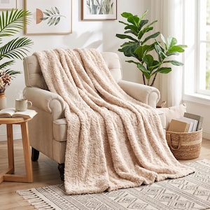 May include: A plush, light beige faux fur throw blanket draped over a cream-colored upholstered armchair. The blanket features a textured, shaggy surface and hangs down from the seat, covering the cushion and backrest of the chair.