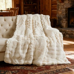 May include: Cream-colored, textured throw blanket draped over a beige sofa. The blanket features a combination of plush, faux fur and a quilted pattern. The cozy blanket is displayed in a rustic living room setting with a fireplace and wooden accents.