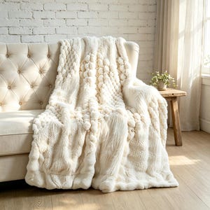 May include: A cream-colored throw blanket featuring a mix of textures, including sections of plush faux fur, raised pom-pom patterns, and intricate cable-knit designs. The blanket is draped over a tufted sofa, showcasing its soft, layered appearance.