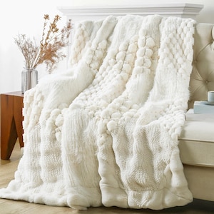 May include: A white faux fur throw blanket with a textured pom-pom design. The blanket is draped over a white sofa.