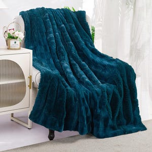 May include: A teal-colored faux fur throw blanket draped over a white armchair. The plush blanket has a textured surface and is folded to show off its soft, inviting appearance. The chair is next to a white cabinet with a gold handle.