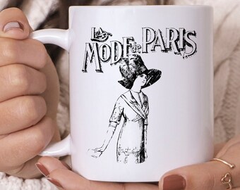May include: White ceramic mug with a black and white illustration of a woman in a hat and the text "La Mode de Paris." The mug has a curved handle and a glossy finish, ideal for tea or coffee.