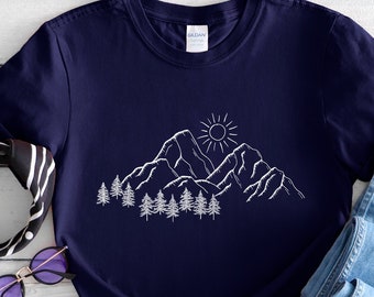 Puede incluir: Una camiseta azul marino con un contorno blanco de montañas, un sol y árboles. La camiseta está colocada sobre una superficie de madera blanca con unas gafas de sol y una bandana blanca y negra.
