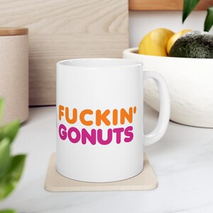 May include: White ceramic mug with the words "FUCKIN' GONUTS" in orange and pink lettering. The mug is on a beige coaster. The mug has a handle and is in front of a wooden cutting board and a bowl of fruit.