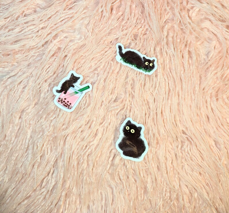 Puede incluir: Tres pegatinas de gatos negros sobre un fondo rosa peludo. Un gato est&aacute; bebiendo t&eacute; de burbujas, otro est&aacute; acostado y el tercero est&aacute; sentado.