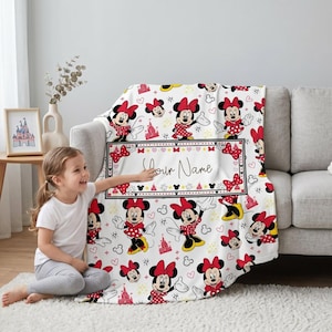 Può includere: Coperta bianca con un motivo ripetuto di Minnie Mouse in rosso e nero. La coperta ha un'area rettangolare con il testo "Your Name" in corsivo, circondata da fiocchi e cuori.