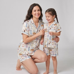 May include: Coordinating pyjama sets for a mother and child. The cream-coloured sets display a repeating pattern of cartoon characters, kitchen tools, and the text "GOSTEAUS". The pyjamas include a collared, button-down shirt and shorts with blue edging.