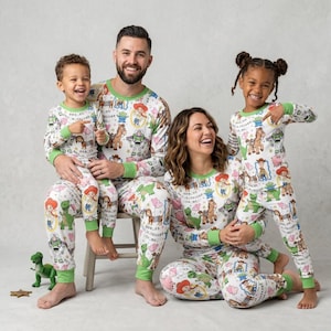 May include: Matching family pajama sets featuring a white background with colorful Toy Story character prints. The long-sleeved tops and pants have green trim. The family of four is smiling and wearing the pajamas.