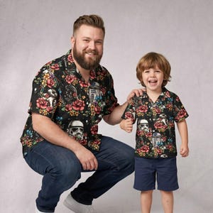 Puede incluir: Dos camisas negras a juego con estampado floral y personajes de Star Wars. Las camisas presentan Stormtroopers y Darth Vader entre flores coloridas. El adulto lleva vaqueros azules y el niño pantalones cortos azul marino.