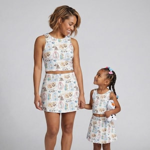 May include: Matching white crop tops and skirts with a colorful cartoon print. The outfits are worn by a woman and a young girl. The girl is holding a stuffed Olaf doll. The print features cartoon characters in pink, blue, and yellow.