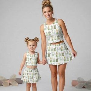 May include: Matching white crop tops and skirts with a whimsical fairy print. The outfits feature a repeating pattern of fairies, potion bottles, and wings in green, yellow, and blue. The outfits are worn by a woman and a young child.