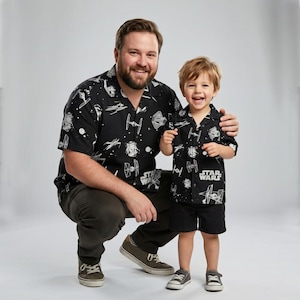 May include: Matching black button-down shirts with a Star Wars theme, featuring white illustrations of spaceships and characters. The shirts are worn by an adult and a child, with the words "STAR WARS" printed on the child's shirt.