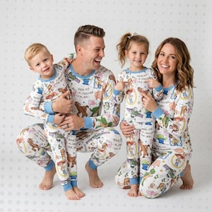 May include: Matching family pajamas featuring a Toy Story theme. The pajamas are white with a colorful print of characters, including Woody, Buzz Lightyear, and others. The long-sleeved tops and pants have blue trim.