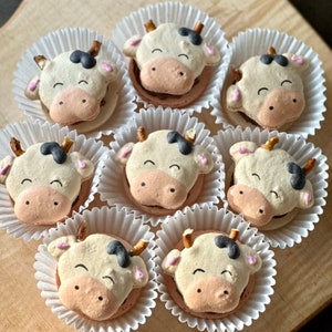 May include: Nine cow-shaped macarons in individual white paper cups. Each macaron is decorated with black spots and a pink nose and mouth.