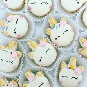 Pode incluir: Uma coleção de macarons com tema de unicórnio, cada um decorado com um chifre dourado, glacê rosa e branco e granulado colorido. Os macarons estão dispostos em forminhas de papel para cupcake brancas.