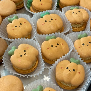 May include: A selection of orange macarons, some shaped as pumpkins with green stems and smiling faces. Other macarons are round. The macarons are in white paper cups, arranged on a tray, and appear to be a seasonal treat.