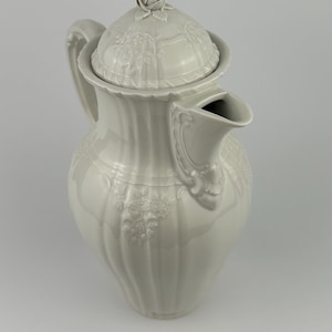 May include: White porcelain coffee pot with a lid and a rose on top. The pot has a decorative embossed design.