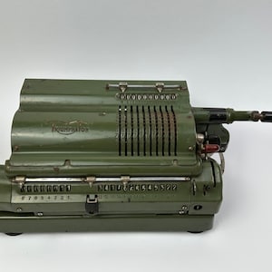 Op de afbeelding: Een groene Triumphator mechanische rekenmachine met een zwarte handgreep en een rij cijferwielen. De rekenmachine heeft een metalen behuizing en is in goede staat.