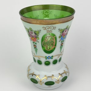 May include: A decorative glass vase with a flared top and a rounded base. The vase features a green rim and accents, with floral and gold detailing. The body is white with green and gold embellishments, and a central green oval.