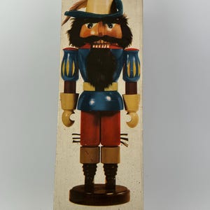 May include: A vintage nutcracker figurine in a box. The nutcracker is dressed in a blue jacket with yellow accents, red pants, and a hat with a feather. The box has a beige background.