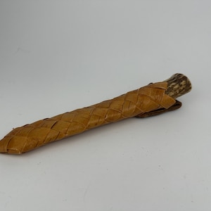 Puede incluir: Un cuchillo artesanal con una funda de cuero trenzado. La funda es de color marrón claro y cubre la hoja, con un mango de asta. El cuchillo está diseñado para uso en exteriores y es un artículo rústico único.