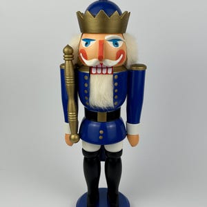 May include: A wooden nutcracker figurine in a royal blue uniform with gold accents. The figurine has a gold crown, a white beard, and a red mouth. It stands on a blue circular base and holds a gold staff. The nutcracker is a classic holiday decoration.