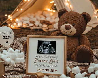 Puede incluir: Un oso de peluche marrón está junto a una imagen de ultrasonido enmarcada. El marco dice "We're adding S'more Love to our family! Baby Miller." Un globo con "Marzo 2024" es visible. Malvaviscos, galletas y una tienda de campaña con luces de hadas están en el fondo.