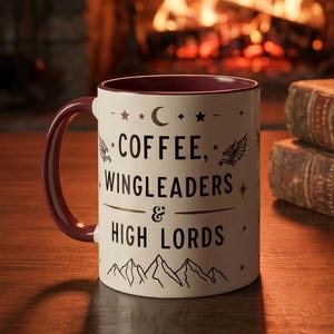 Puede incluir: Una taza de cerámica blanca con interior y asa burdeos, con el texto "COFFEE, WINGLEADERS & HIGH LORDS" en negro. La taza está decorada con estrellas, una luna creciente y gráficos de montañas. Una pluma descansa sobre una pila de libros antiguos.