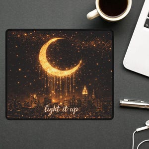 May include: Black rectangular mousepad featuring a golden crescent moon with sparkling accents and a cityscape silhouette. The words "light it up" are printed below the city. A cup of coffee and a laptop are in the background.