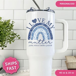 May include: A white stainless steel tumbler with a handle and a lid. The front features a blue floral patterned rainbow graphic with the text "I LOVE MY mutter" and the names "JANE NICK EMMA JASON" printed in blue below the rainbow.
