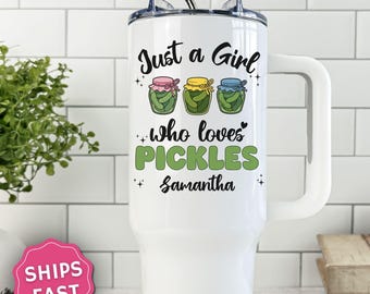 May include: A white stainless steel tumbler with a handle and a straw, featuring the text "Just a Girl who loves PICKLES" and the name "Samantha" in black and green, accompanied by illustrations of three pickle jars with pink, yellow, and blue lids.